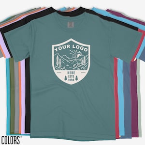 K&ouml;nnte beinhalten: Eine Reihe von Comfort Colors T-Shirts in verschiedenen Farben, darunter T&uuml;rkis, Schwarz und Lila. Das vordere Shirt zeigt eine wei&szlig;e Grafik mit dem Text "YOUR LOGO" und einer Berglandschaft. Der Text "HERE ESTD 1998" ist ebenfalls sichtbar.
