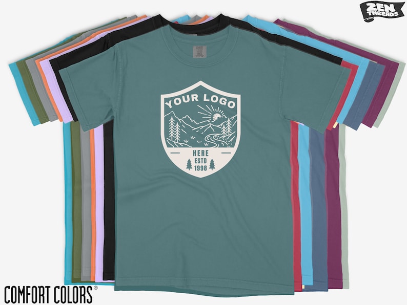 K&ouml;nnte beinhalten: Eine Reihe von Comfort Colors T-Shirts in verschiedenen Farben, darunter T&uuml;rkis, Schwarz und Lila. Das vordere Shirt zeigt eine wei&szlig;e Grafik mit dem Text "YOUR LOGO" und einer Berglandschaft. Der Text "HERE ESTD 1998" ist ebenfalls sichtbar.