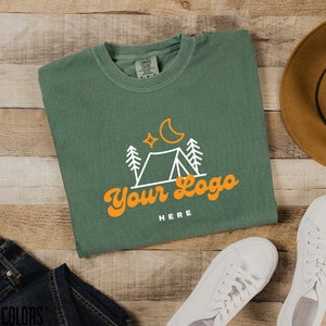 May include: A green t-shirt featuring a white outline of a tent, trees, a crescent moon, and stars. The text "Your Logo" is in orange with the words "HERE" underneath.