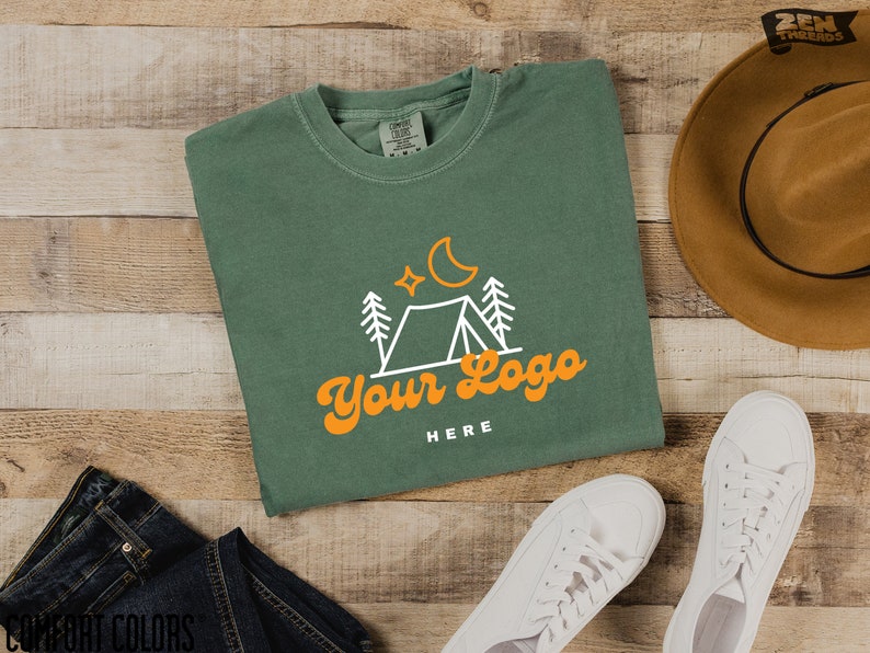 May include: A green t-shirt featuring a white outline of a tent, trees, a crescent moon, and stars. The text "Your Logo" is in orange with the words "HERE" underneath.