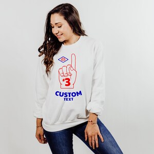 Puede incluir: Sudadera blanca con un dise&ntilde;o rojo y azul de un dedo de espuma apuntando hacia arriba, con el n&uacute;mero 3 y las palabras "CUSTOM TEXT". Un peque&ntilde;o logotipo azul y rojo en forma de diamante est&aacute; encima del dedo. La sudadera la lleva una persona.
