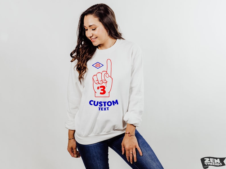 Puede incluir: Sudadera blanca con un dise&ntilde;o rojo y azul de un dedo de espuma apuntando hacia arriba, con el n&uacute;mero 3 y las palabras "CUSTOM TEXT". Un peque&ntilde;o logotipo azul y rojo en forma de diamante est&aacute; encima del dedo. La sudadera la lleva una persona.