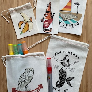 Op de afbeelding: Vijf witte katoenen trekkoordtassen met verschillende bedrukte ontwerpen. De ontwerpen omvatten een banaan, een bierfles, een vlot met een zeil, een zeemeermin en een uil. De tassen liggen op een houten oppervlak met vier kleurpotloden en een rode stift.