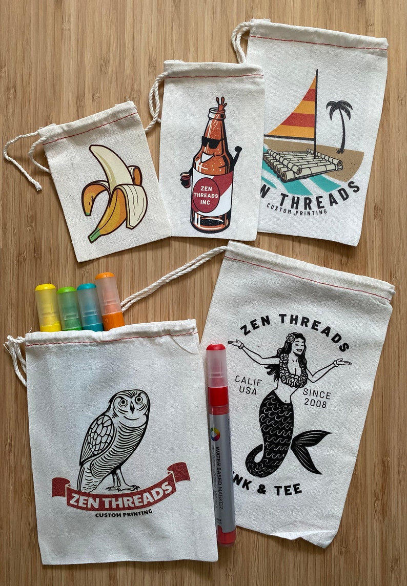 Op de afbeelding: Vijf witte katoenen trekkoordtassen met verschillende bedrukte ontwerpen. De ontwerpen omvatten een banaan, een bierfles, een vlot met een zeil, een zeemeermin en een uil. De tassen liggen op een houten oppervlak met vier kleurpotloden en een rode stift.