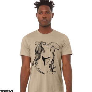 May include: A tan t-shirt with a black and white illustration of a hammerhead shark, a shark tooth, and a shark fin.