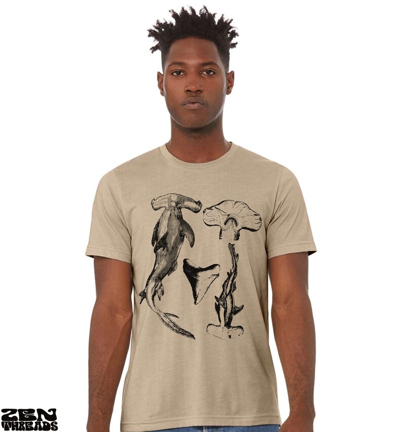 May include: A tan t-shirt with a black and white illustration of a hammerhead shark, a shark tooth, and a shark fin.