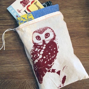 May include: A white canvas bag with a drawstring closure, featuring a red and white owl print. The bag is filled with various items, including postcards and a blue and yellow patterned fabric.
