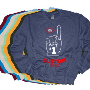 Puede incluir: Una sudadera azul marino con un gr&aacute;fico de mano blanca que apunta hacia arriba, el n&uacute;mero 1 y las palabras "CUSTOM TEXT" en rojo. En la esquina superior izquierda hay un peque&ntilde;o diamante con las palabras "YOUR ART OR PHOTO". Varias otras sudaderas de diferentes colores est&aacute;n apiladas detr&aacute;s.