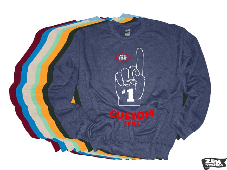 Puede incluir: Una sudadera azul marino con un gr&aacute;fico de mano blanca que apunta hacia arriba, el n&uacute;mero 1 y las palabras "CUSTOM TEXT" en rojo. En la esquina superior izquierda hay un peque&ntilde;o diamante con las palabras "YOUR ART OR PHOTO". Varias otras sudaderas de diferentes colores est&aacute;n apiladas detr&aacute;s.
