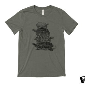 May include: A heather green t-shirt with a black and white illustration of a stack of turtles. The design shows three turtles stacked on top of each other. The shirt has a crew neck and short sleeves.