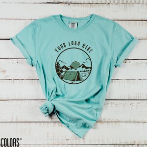 K&ouml;nnte beinhalten: T&uuml;rkisfarbenes Comfort Colors T-Shirt mit einem kreisf&ouml;rmigen Grafikdesign, das ein Zelt, Berge und den Text "YOUR LOGO HERE" zeigt. Das T-Shirt hat hochgekrempelte &Auml;rmel und wird auf einer wei&szlig;en Holzoberfl&auml;che pr&auml;sentiert. Das Zen Threads-Logo befindet sich oben rechts.