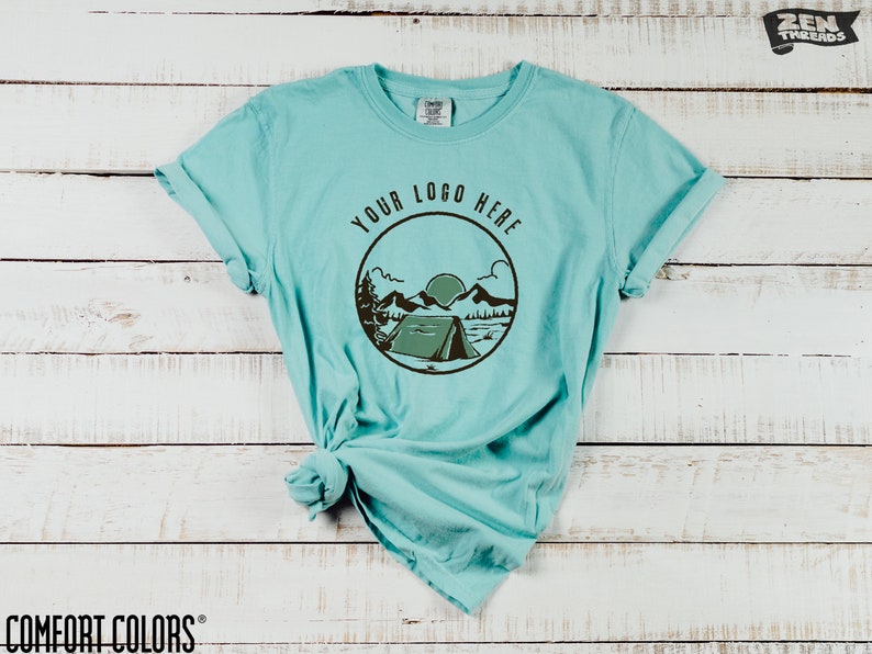 K&ouml;nnte beinhalten: T&uuml;rkisfarbenes Comfort Colors T-Shirt mit einem kreisf&ouml;rmigen Grafikdesign, das ein Zelt, Berge und den Text "YOUR LOGO HERE" zeigt. Das T-Shirt hat hochgekrempelte &Auml;rmel und wird auf einer wei&szlig;en Holzoberfl&auml;che pr&auml;sentiert. Das Zen Threads-Logo befindet sich oben rechts.