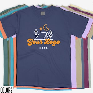 May include: A navy blue t-shirt featuring a white camping scene and the text "Your Logo Here" in orange. The shirt is part of a stack of t-shirts in various colours.