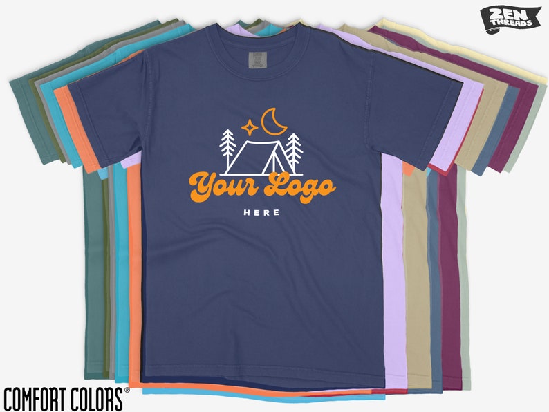 May include: A navy blue t-shirt featuring a white camping scene and the text "Your Logo Here" in orange. The shirt is part of a stack of t-shirts in various colours.