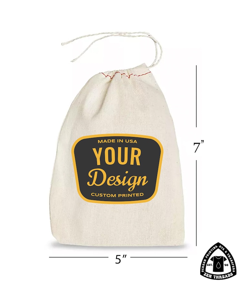 Op de afbeelding: Een witte katoenen trekkoordtas met een zwart en geel label waarop staat "Made in USA, Your Design, Custom Printed". De tas is 17,8 cm hoog en 12,7 cm breed.