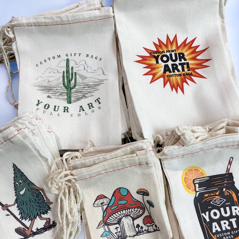 Op de afbeelding: Een stapel witte katoenen trekkoordtassen met verschillende ontwerpen erop gedrukt. De ontwerpen omvatten een cactus, een paddenstoel, een citroen en een glazen pot. De tekst op de tassen luidt "Custom Gift Bags", "Your Art", "Custom Print" en "Cotton Bags".