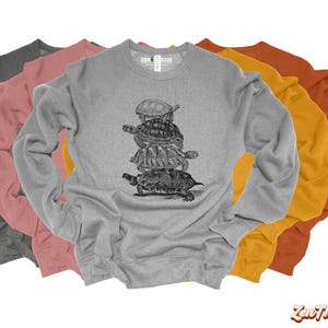 May include: A gray sweatshirt with a graphic of four stacked turtles. The sweatshirt is shown with three other sweatshirts in pink, yellow, and orange.