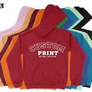 May include: A stack of red, orange, maroon, yellow, lavender, green, blue, teal, black, and light green hooded sweatshirts. The front of the red sweatshirt has white text that reads "CUSTOM PRINT ANY COLOR - TEXT OR LOGO".