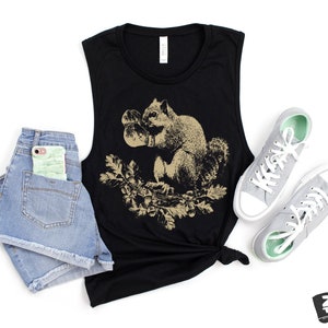 May include: A black sleeveless muscle tank top with a graphic print of a squirrel holding an acorn. The squirrel is in a beige color and the background is black. The tank top is laying on a white surface with a pair of gray canvas shoes and a pair of denim shorts.