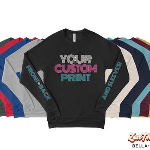May include: A black long-sleeve raglan shirt with the text "YOUR CUSTOM PRINT" in white, pink, and teal. The sleeves have the text "FRONT BACK" and "AND SLEEVES!". Several other long-sleeve shirts in various colors are behind the black shirt.