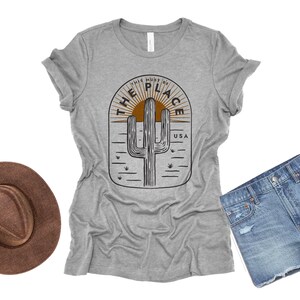 May include: A heather gray t-shirt with a desert-themed graphic featuring a cactus, sun, and the text "THIS MUST BE THE PLACE USA." A brown hat and denim shorts are also in the image.