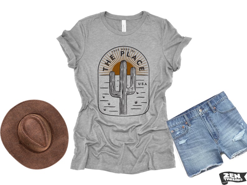 May include: A heather gray t-shirt with a desert-themed graphic featuring a cactus, sun, and the text "THIS MUST BE THE PLACE USA." A brown hat and denim shorts are also in the image.