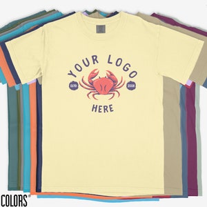 May include: A stack of t-shirts in various colors, including teal, olive green, orange, navy blue, lavender, beige, and maroon. The top shirt is yellow with a red crab graphic and the text "YOUR LOGO" in a circle with "ESTD 2008".