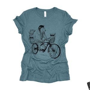 May include: A teal blue t-shirt with a black and white illustration of a bear riding a bicycle with a basket full of books.