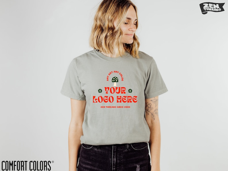 K&ouml;nnte beinhalten: Salbeigr&uuml;nes T-Shirt mit dem Text "YOUR LOGO HERE" in Rot, mit einem Pilz-Motiv. Das Shirt hat auch den Text "ANY ART ANY COLOR" und "ZEN THREADS SINCE 2000". Das Shirt ist von Comfort Colors.