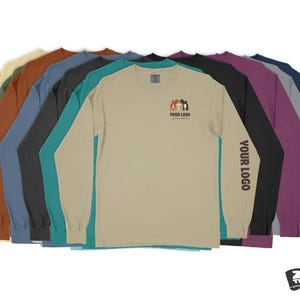 Pode incluir: Uma sele&ccedil;&atilde;o de camisetas de manga comprida em v&aacute;rias cores, incluindo bege, marrom, azul-turquesa, azul e roxo. A camiseta bege apresenta um pequeno gr&aacute;fico de uma casa com o texto "YOUR LOGO" abaixo. A camiseta roxa tem o texto "YOUR LOGO" impresso na manga.