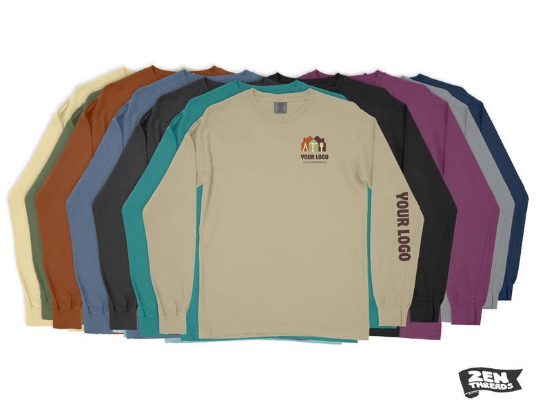 Pode incluir: Uma sele&ccedil;&atilde;o de camisetas de manga comprida em v&aacute;rias cores, incluindo bege, marrom, azul-turquesa, azul e roxo. A camiseta bege apresenta um pequeno gr&aacute;fico de uma casa com o texto "YOUR LOGO" abaixo. A camiseta roxa tem o texto "YOUR LOGO" impresso na manga.