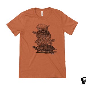 May include: A rust-coloured t-shirt with a black and white illustration of four turtles piled on top of each other. The shirt has a crew neck and short sleeves. The brand logo "ZEN THREADS" is in the bottom right corner.