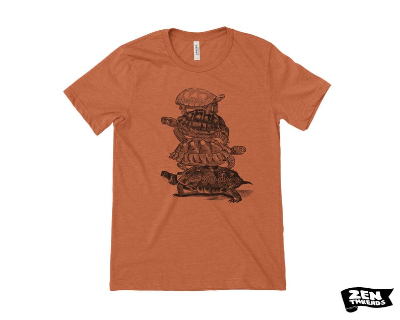 May include: A rust-coloured t-shirt with a black and white illustration of four turtles piled on top of each other. The shirt has a crew neck and short sleeves. The brand logo "ZEN THREADS" is in the bottom right corner.