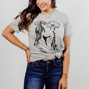 May include: A heather grey t-shirt with a graphic print of a hammerhead shark, a shark tooth, and a stingray. The shirt is tied at the waist.