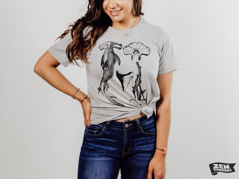 May include: A heather grey t-shirt with a graphic print of a hammerhead shark, a shark tooth, and a stingray. The shirt is tied at the waist.