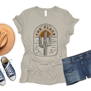 May include: A light gray t-shirt with a desert-themed graphic, denim shorts, a brown hat, and blue sneakers. The t-shirt features a cactus design with the text "THIS MUST BE THE PLACE USA".