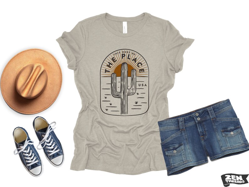 May include: A light gray t-shirt with a desert-themed graphic, denim shorts, a brown hat, and blue sneakers. The t-shirt features a cactus design with the text "THIS MUST BE THE PLACE USA".