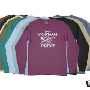 Pode incluir: Uma pilha de camisas de manga comprida em v&aacute;rias cores, incluindo marrom, bege, azul-turquesa, azul claro, rosa e preto. A camisa da frente &eacute; roxa com um gr&aacute;fico branco que diz "Your Custom Print" e um endere&ccedil;o de site.