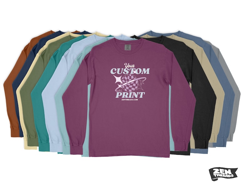 Pode incluir: Uma pilha de camisas de manga comprida em v&aacute;rias cores, incluindo marrom, bege, azul-turquesa, azul claro, rosa e preto. A camisa da frente &eacute; roxa com um gr&aacute;fico branco que diz "Your Custom Print" e um endere&ccedil;o de site.