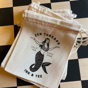 Op de afbeelding: Een stapel van drie witte katoenen trekkoordtassen met rode stiksels. De tassen hebben een zwart-wit logo met de tekst "ZEN THREADS", "CALIF. USA", "SINCE 2008" en "INK & TEE". Het logo bevat ook een zwart-witte illustratie van een zeemeermin.