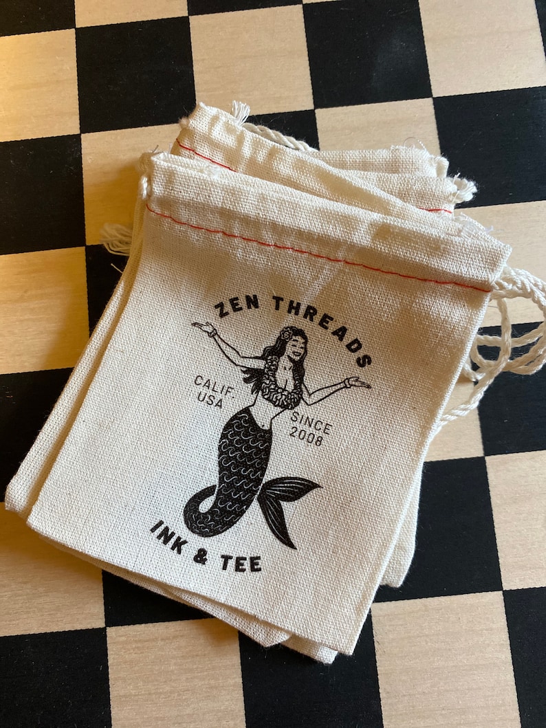 Op de afbeelding: Een stapel van drie witte katoenen trekkoordtassen met rode stiksels. De tassen hebben een zwart-wit logo met de tekst "ZEN THREADS", "CALIF. USA", "SINCE 2008" en "INK & TEE". Het logo bevat ook een zwart-witte illustratie van een zeemeermin.