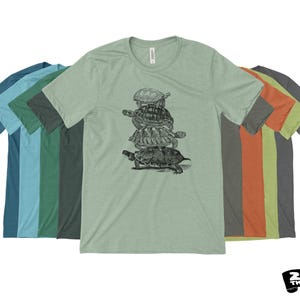 May include: A light green crew neck t-shirt featuring a black and white illustration of several turtles stacked on top of each other. The shirt is surrounded by other t-shirts in various colors, including blue, green, and orange. The Zen Threads logo is in the bottom right corner.