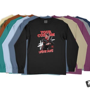 Pode incluir: Uma camiseta preta de manga comprida com um gr&aacute;fico vermelho e branco de um jogador de futebol americano. O texto "YOUR CUSTOM LOGO OR IMAGE" &eacute; impresso abaixo do gr&aacute;fico. A camiseta &eacute; mostrada com uma variedade de op&ccedil;&otilde;es de cores.