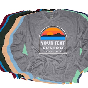 Puede incluir: Una sudadera gris de manga larga con un dise&ntilde;o personalizado. El dise&ntilde;o presenta una puesta de sol sobre monta&ntilde;as con el texto "YOUR TEXT CUSTOM" y "LOGO OR PHOTO". Se ven varias sudaderas de otros colores.