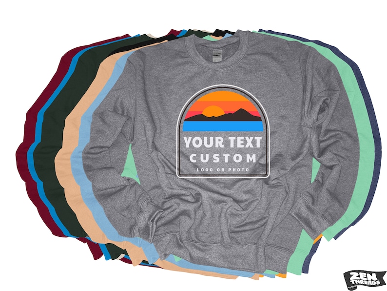 Puede incluir: Una sudadera gris de manga larga con un dise&ntilde;o personalizado. El dise&ntilde;o presenta una puesta de sol sobre monta&ntilde;as con el texto "YOUR TEXT CUSTOM" y "LOGO OR PHOTO". Se ven varias sudaderas de otros colores.