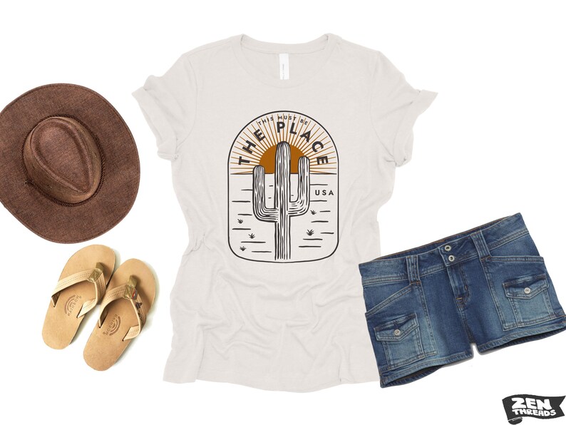 May include: A white t-shirt with a desert-themed graphic, featuring a cactus and the text "THIS MUST BE THE PLACE USA." The image also includes a brown hat, denim shorts, and tan sandals, suggesting a casual, outdoor theme.