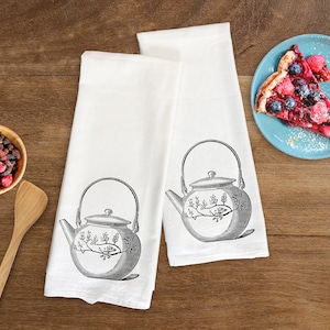 May include: Two white tea towels with a black and white illustration of a teapot with floral designs. The tea towels are on a wooden surface with a plate of fruit and a wooden spoon.