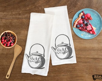 May include: Two white tea towels with a black and white illustration of a teapot with floral designs. The tea towels are on a wooden surface with a plate of fruit and a wooden spoon.