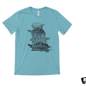 May include: A light blue t-shirt with a black and white illustration of turtles stacked on top of each other. The shirt has a crew neck and short sleeves. The brand name "ZEN THREADS" is printed in black on a white background.