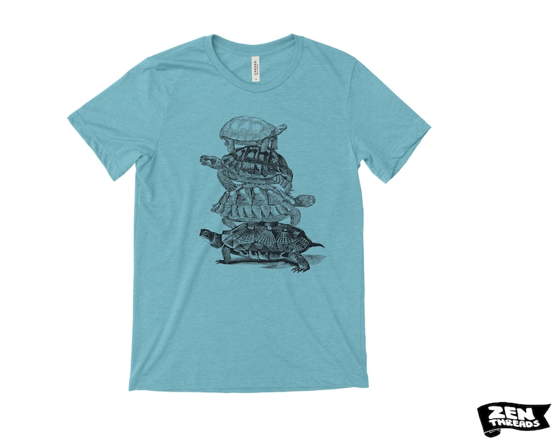 May include: A light blue t-shirt with a black and white illustration of turtles stacked on top of each other. The shirt has a crew neck and short sleeves. The brand name "ZEN THREADS" is printed in black on a white background.